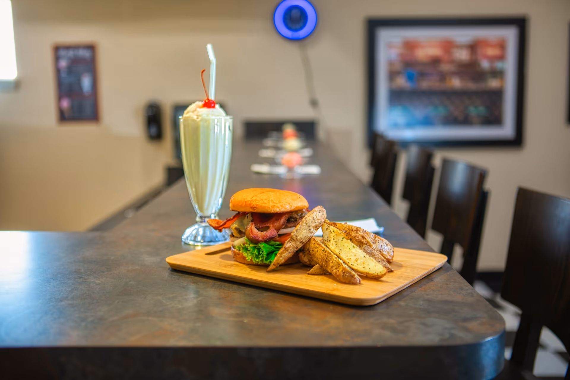A wooden tray on a countertop holding a bacon cheeseburger with lettuce and tomato, accompanied by seasoned potato wedges. Next to the tray is a tall glass of vanilla milkshake topped with whipped cream and a cherry, with a straw. The background shows a row of chairs along the counter and framed pictures on the wall.