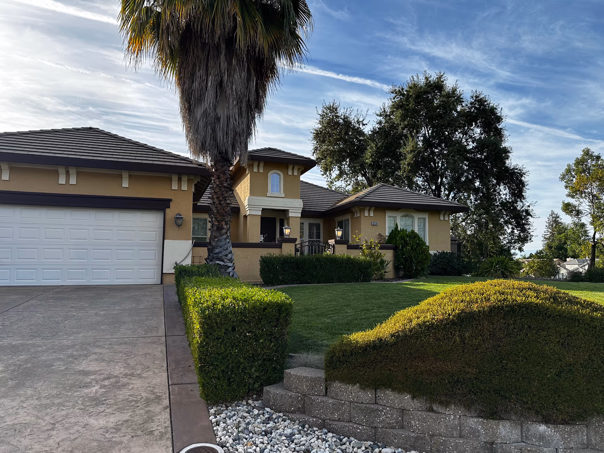 Exterior view of a single-story house with a beige facade, a two-car garage with a white door, a tall palm tree in front, neatly trimmed hedges, a green lawn, and a partly cloudy sky.