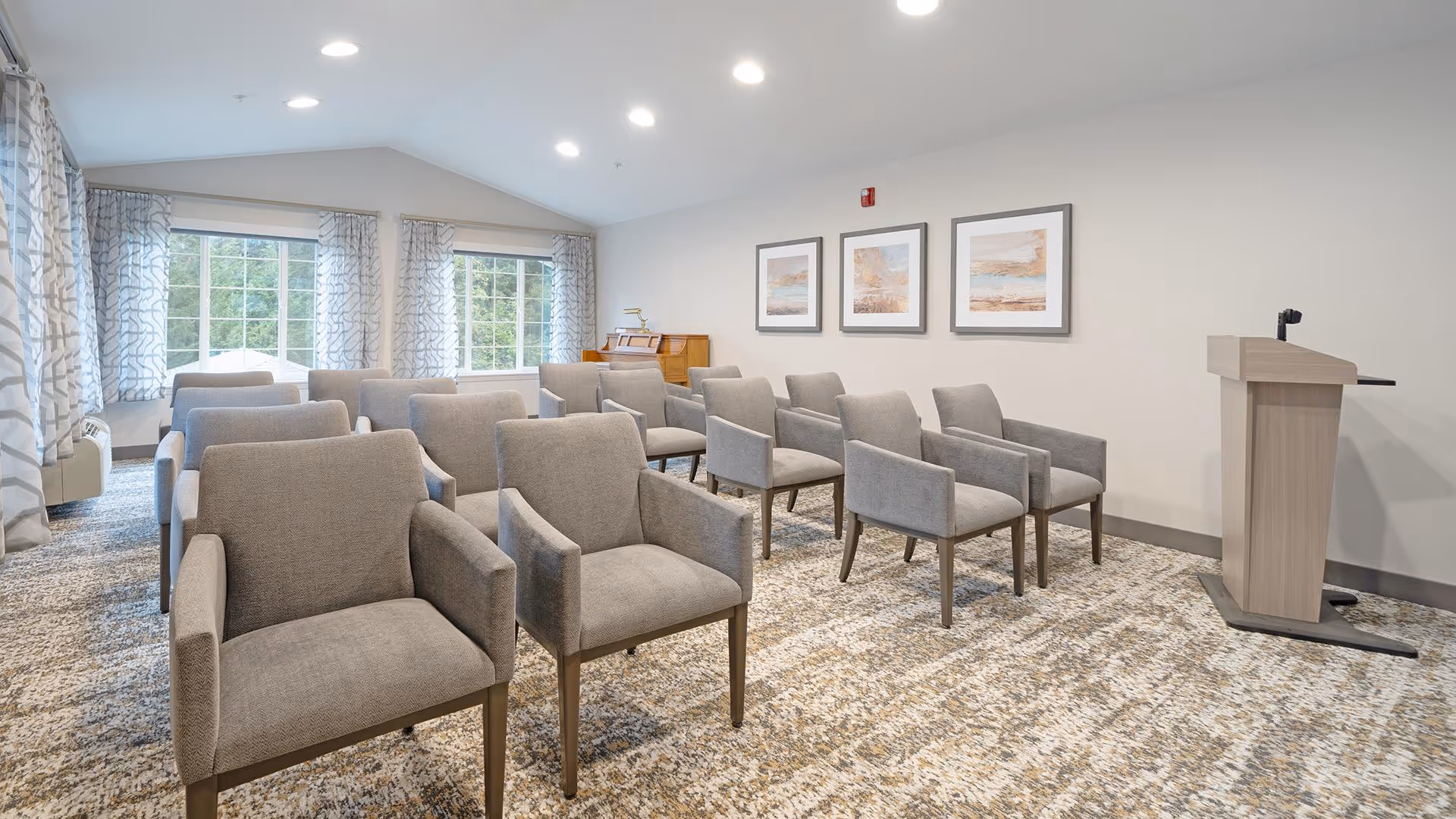 A well-lit room with rows of gray upholstered chairs arranged facing a wooden podium with a microphone. The room has large windows with patterned curtains, three framed artworks on the wall, and a carpeted floor with a textured pattern.