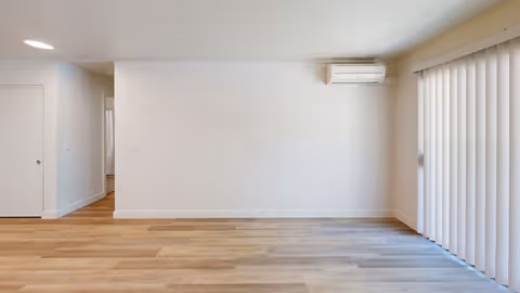 Empty room with light wood flooring, white walls, a white door on the left, a wall-mounted air conditioning unit, and vertical blinds covering a window on the right.
