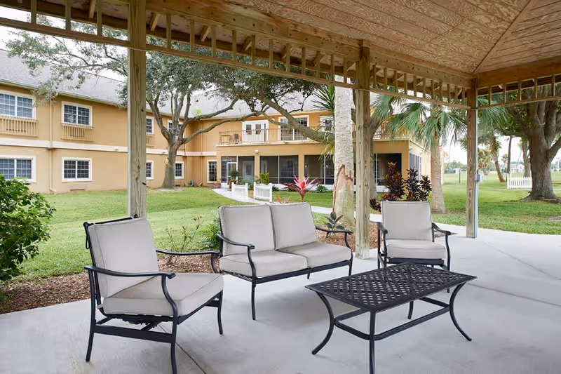 Outdoor covered patio area with cushioned metal chairs and a matching coffee table, overlooking a grassy lawn and a two-story beige building with balconies and windows, surrounded by trees and plants.