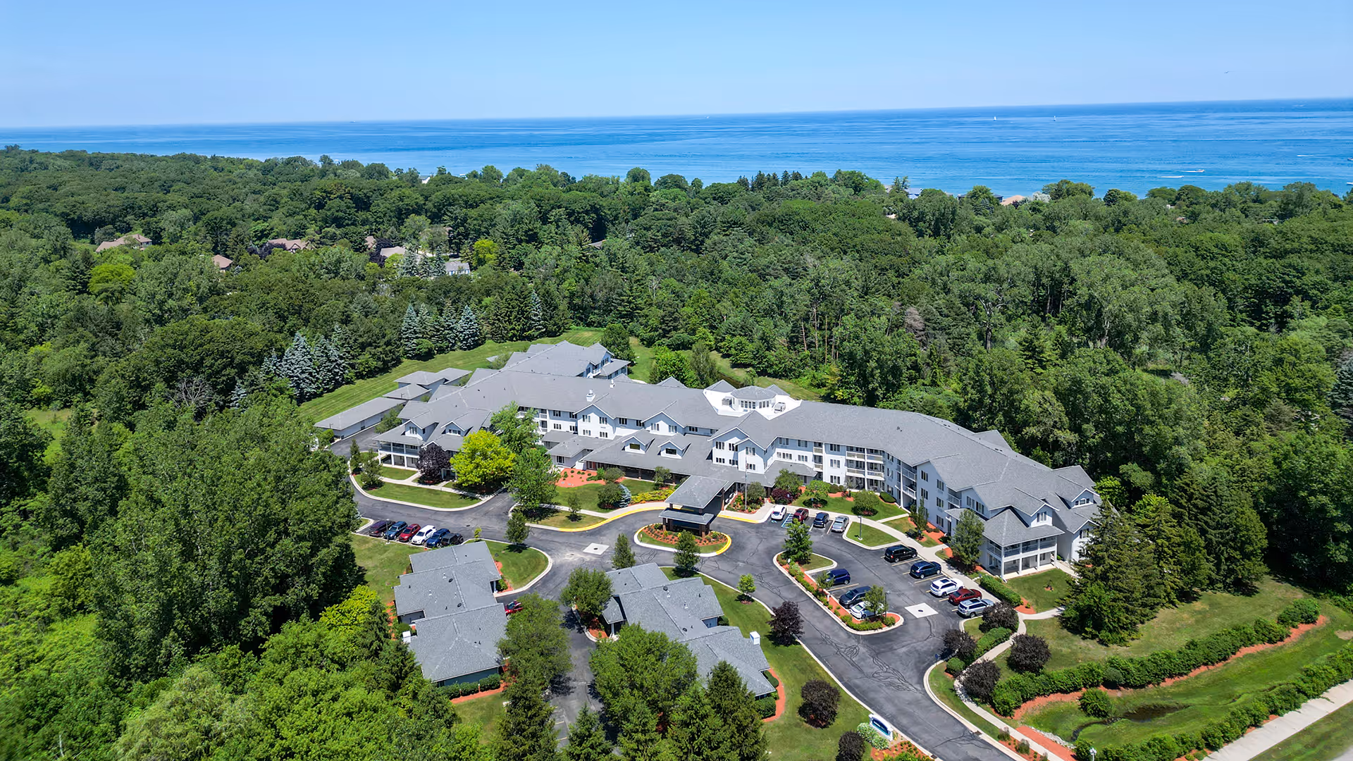 Aerial view of Holiday Blue Water Lodge surrounded by dense green trees with a large body of water visible in the background. The facility consists of multiple connected buildings with gray roofs, parking areas with cars, and landscaped grounds.