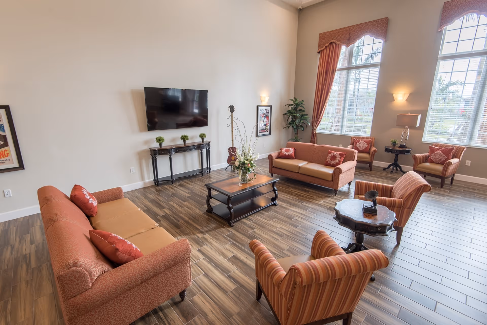 A spacious living room with wooden flooring, two orange sofas, two striped armchairs, a coffee table with a flower arrangement, a side table with a lamp, a flat-screen TV mounted on the wall, large windows with red curtains, and framed artwork on the walls.