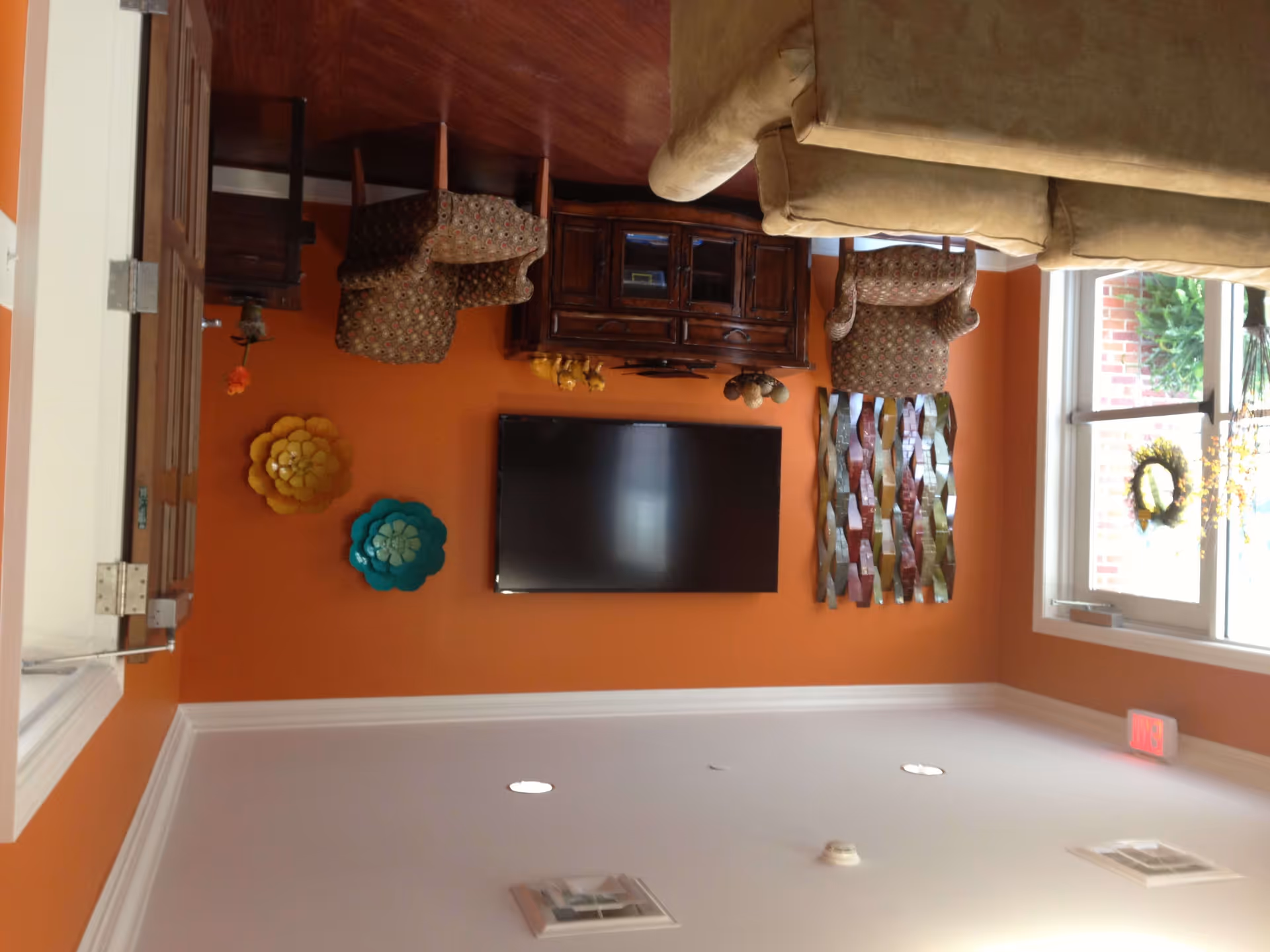A cozy living room with orange walls featuring a mounted flat-screen TV, two patterned armchairs, a beige sofa, a wooden cabinet, and decorative wall art including colorful flower-shaped pieces and a metal sculpture. A window with a wreath allows natural light into the room.