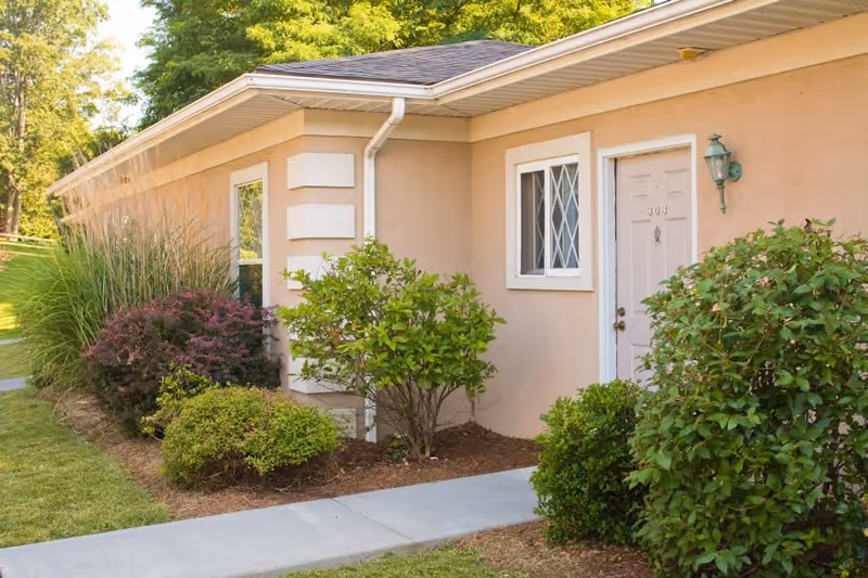 Exterior view of a beige single-story building with a white door numbered 303, a window with white framing, a green outdoor wall lamp, and various green shrubs and plants along a concrete walkway.