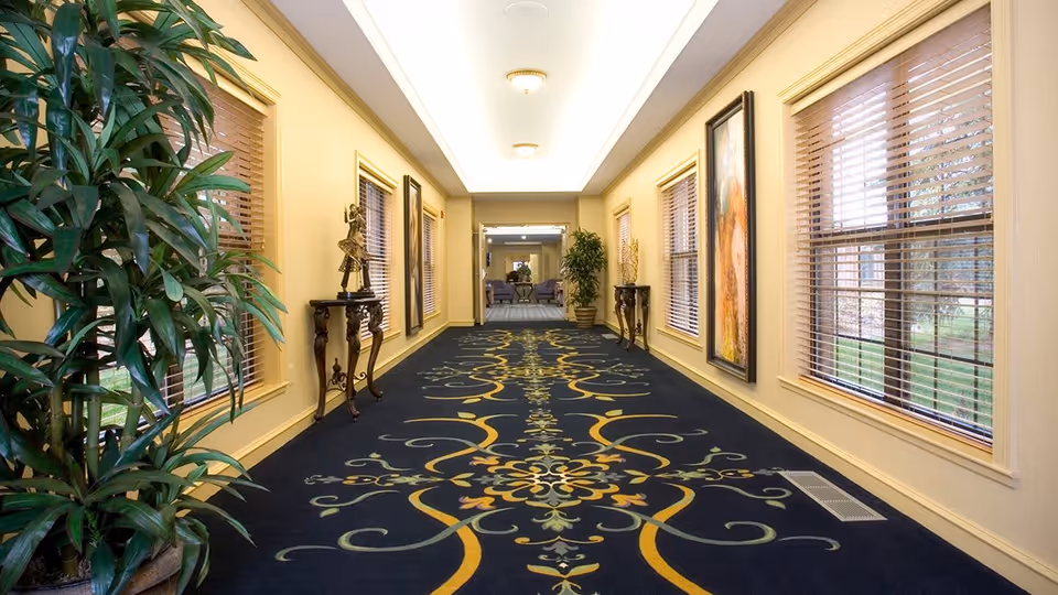 A long, well-lit hallway with large windows on the right side covered with wooden blinds. The hallway has a dark carpet with an intricate yellow and green floral pattern. There are decorative tables with sculptures and plants along the left wall, and framed artwork on the right wall. At the end of the hallway, there is a seating area with chairs and a table.