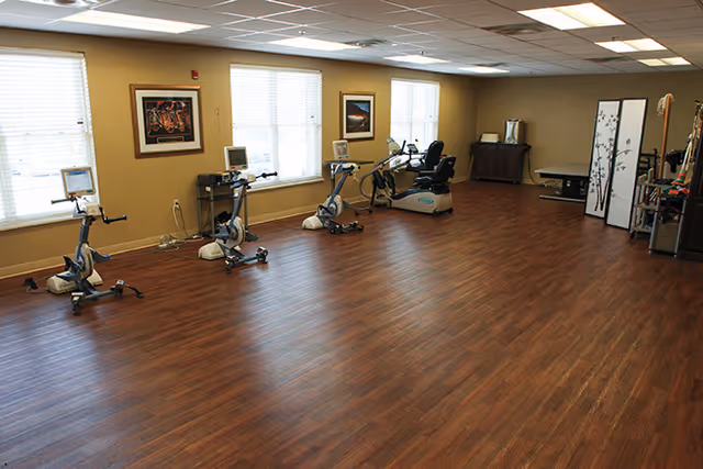 A spacious room with wooden flooring featuring several exercise bikes lined up against a wall with three windows. The room has beige walls with framed pictures, a wooden cabinet, a folding screen, and some exercise equipment in the corner.