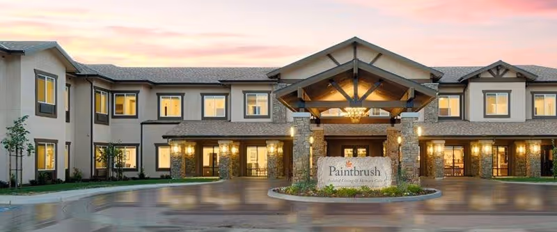Exterior view of Paintbrush Assisted Living facility at dusk, showing a two-story building with warm interior lights, a covered entrance with stone pillars, and a sign reading 'Paintbrush Assisted Living of Missoula, Montana' in front.