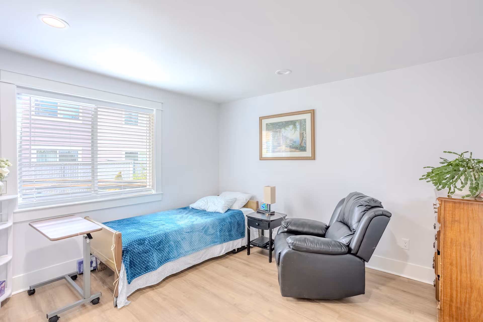 Bright single-occupancy bedroom with a bed covered by a blue blanket, a black recliner, side table, dresser and a large window with blinds.