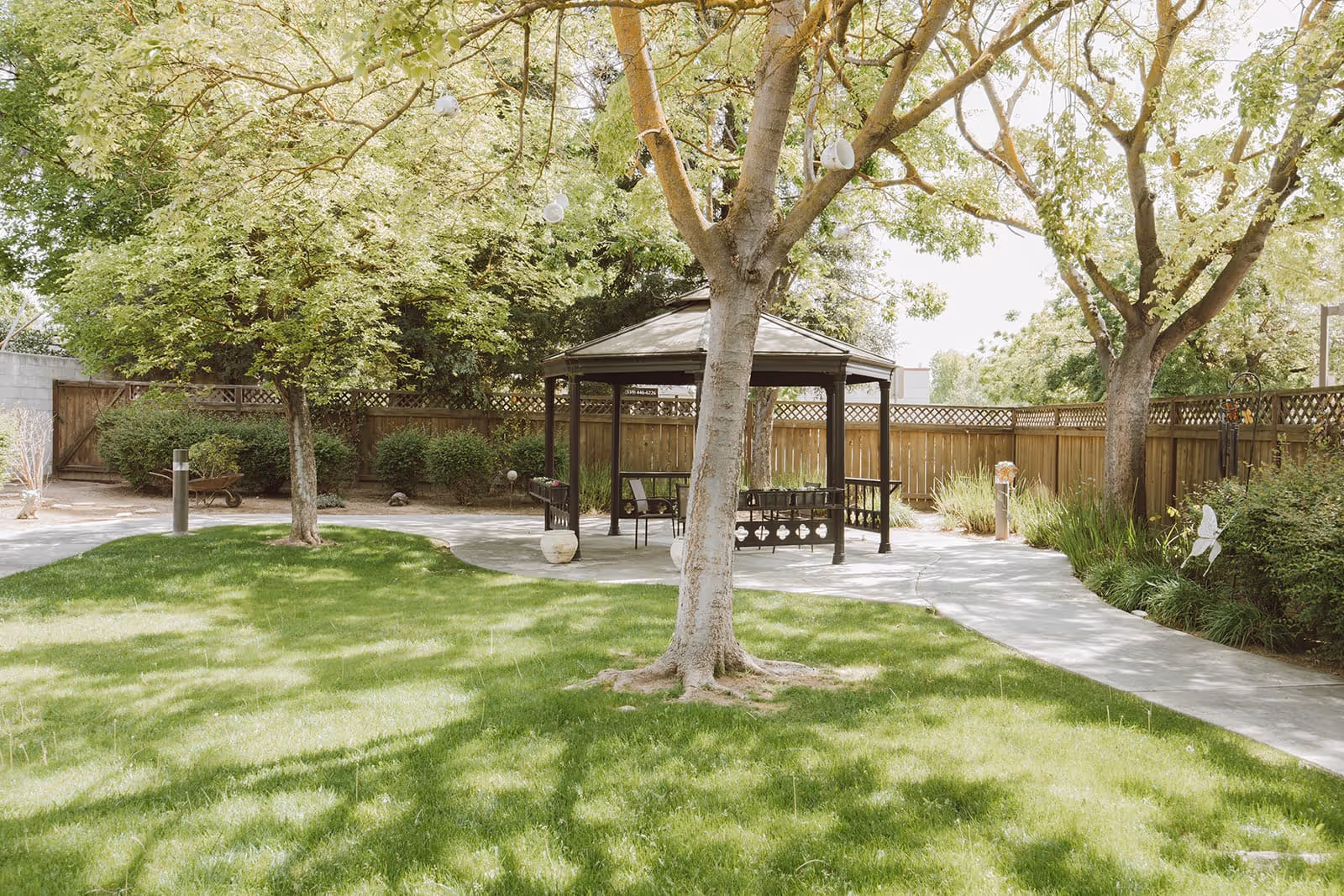 A peaceful outdoor garden area with green grass, several trees, and a wooden gazebo with benches underneath. A paved walkway curves through the garden, and a wooden fence encloses the space.