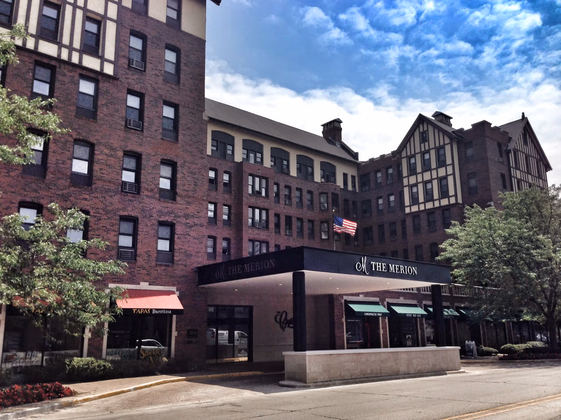 Exterior view of The Merion building, a multi-story brick structure with Tudor-style architectural elements. The entrance features a covered driveway with a sign displaying 'The Merion' and an American flag flying above. Trees and a sidewalk line the street in front of the building.
