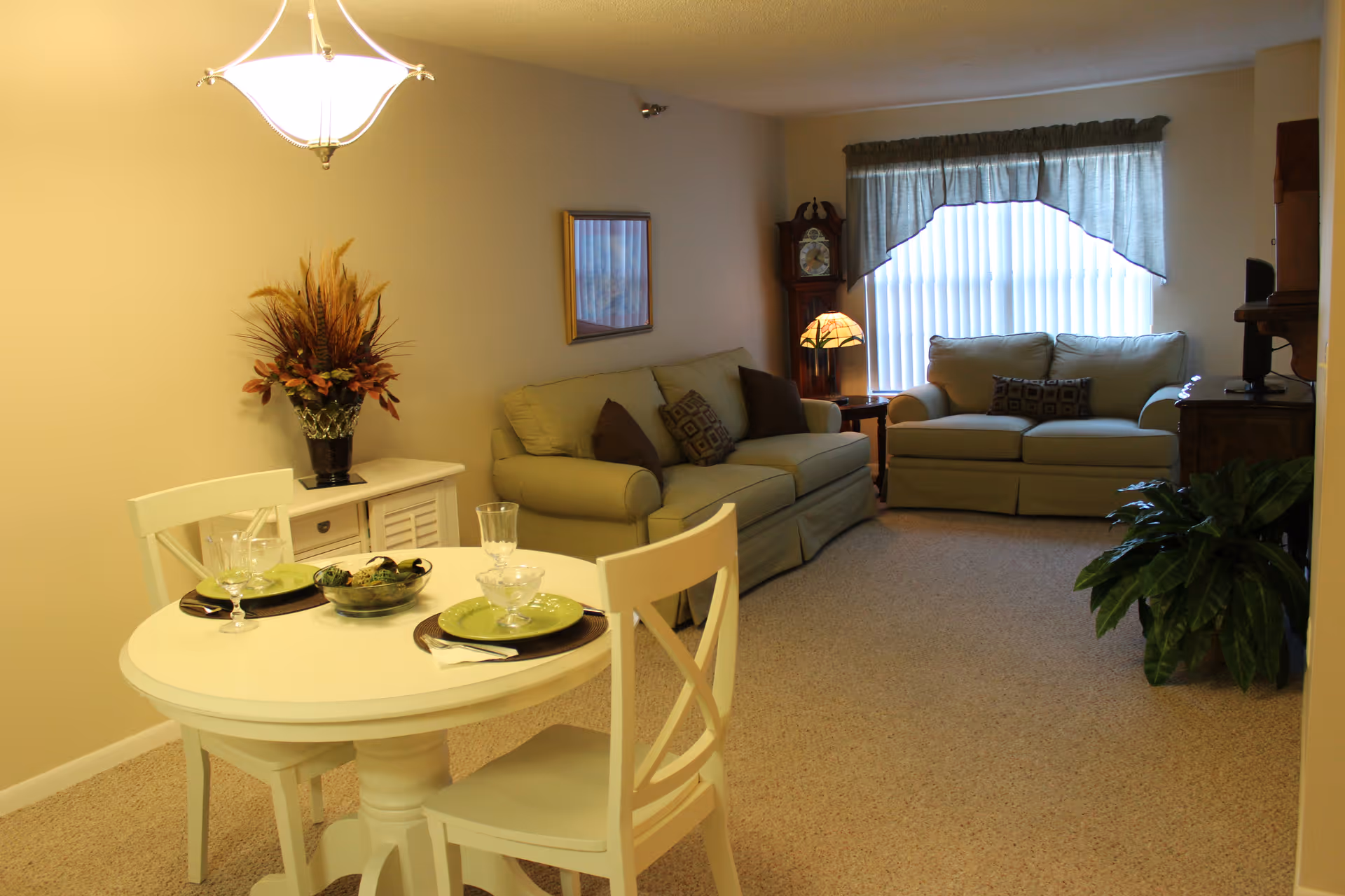 A furnished living and dining area with a small round table set for two in the foreground and sofas, a lamp, and a window with vertical blinds in the background.