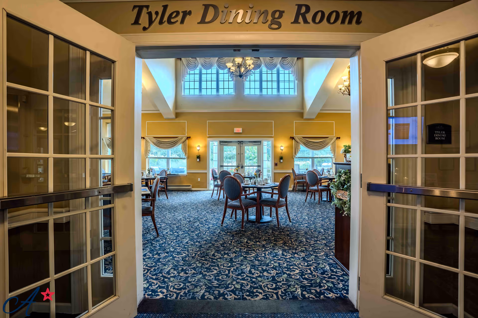 View through open double doors into the Tyler Dining Room, featuring a carpeted floor with a blue and white pattern, round dining tables with chairs, large windows letting in natural light, and chandeliers hanging from a high ceiling.
