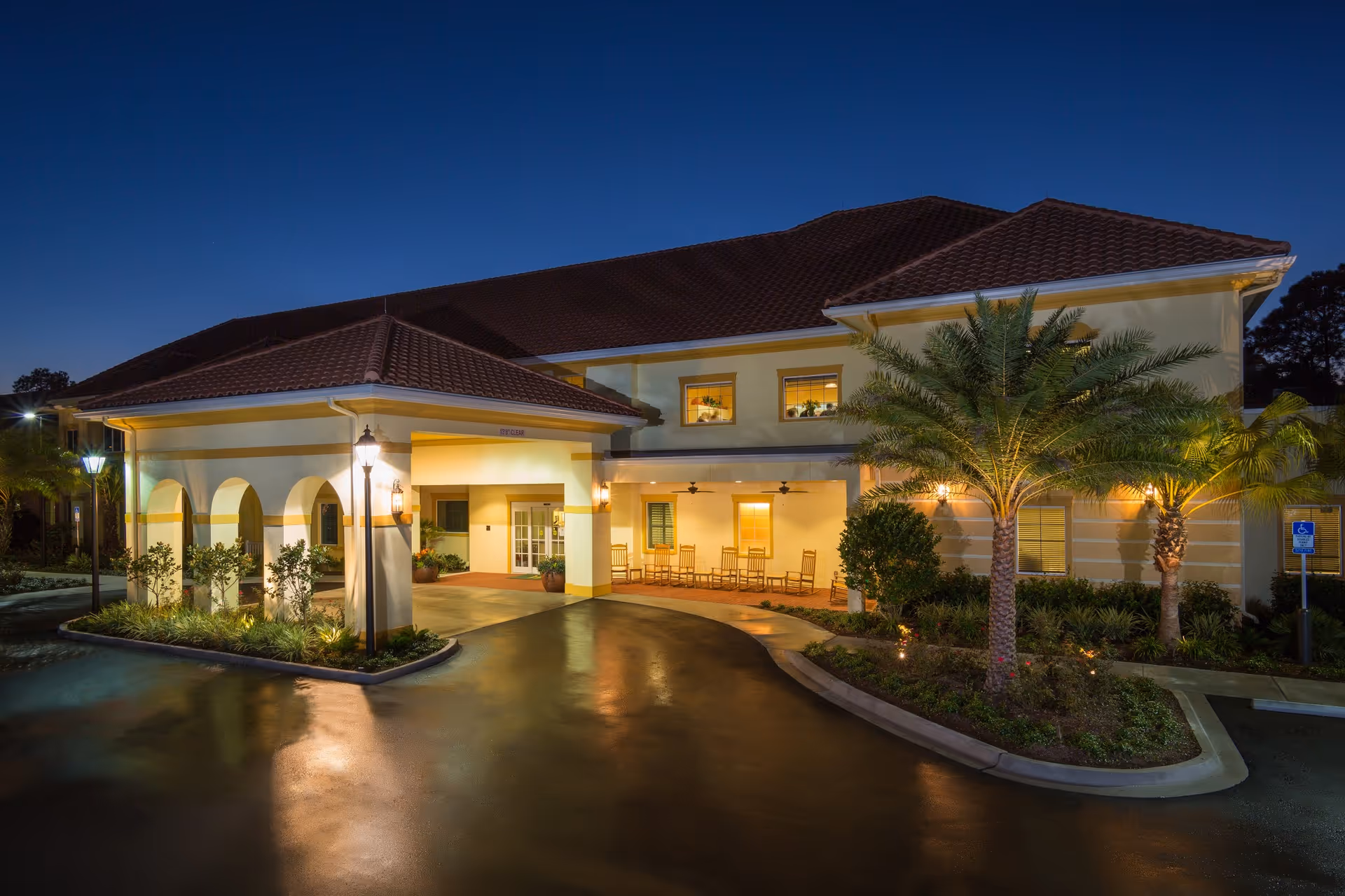 Exterior view of The Windsor at Ortega senior living facility at dusk, showing a well-lit entrance with a covered driveway, palm trees, and rocking chairs on the porch.