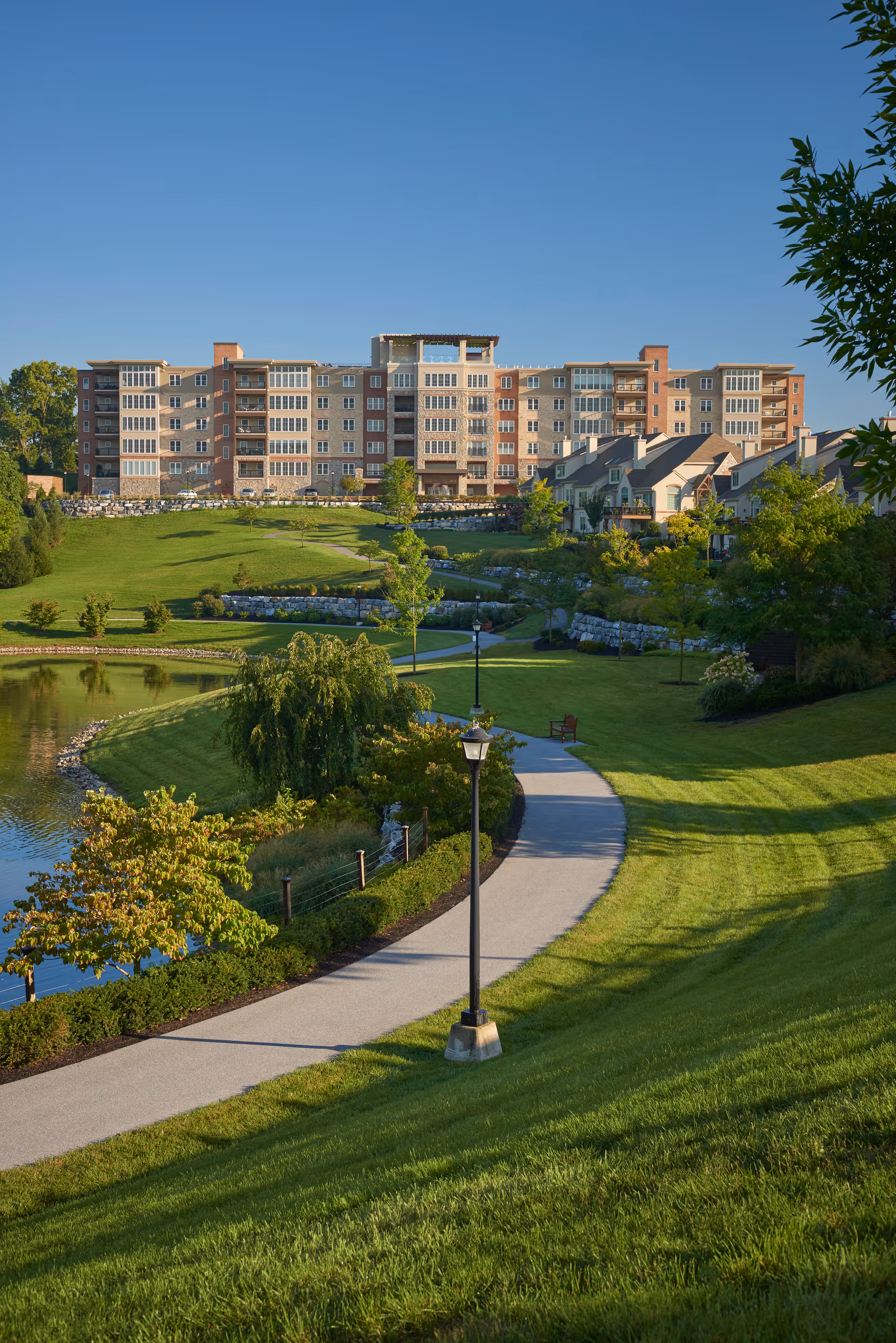 A scenic outdoor view of The Vistas at Willow Valley Communities featuring a large multi-story residential building in the background, surrounded by well-maintained green lawns, trees, a walking path, and a small pond under a clear blue sky.