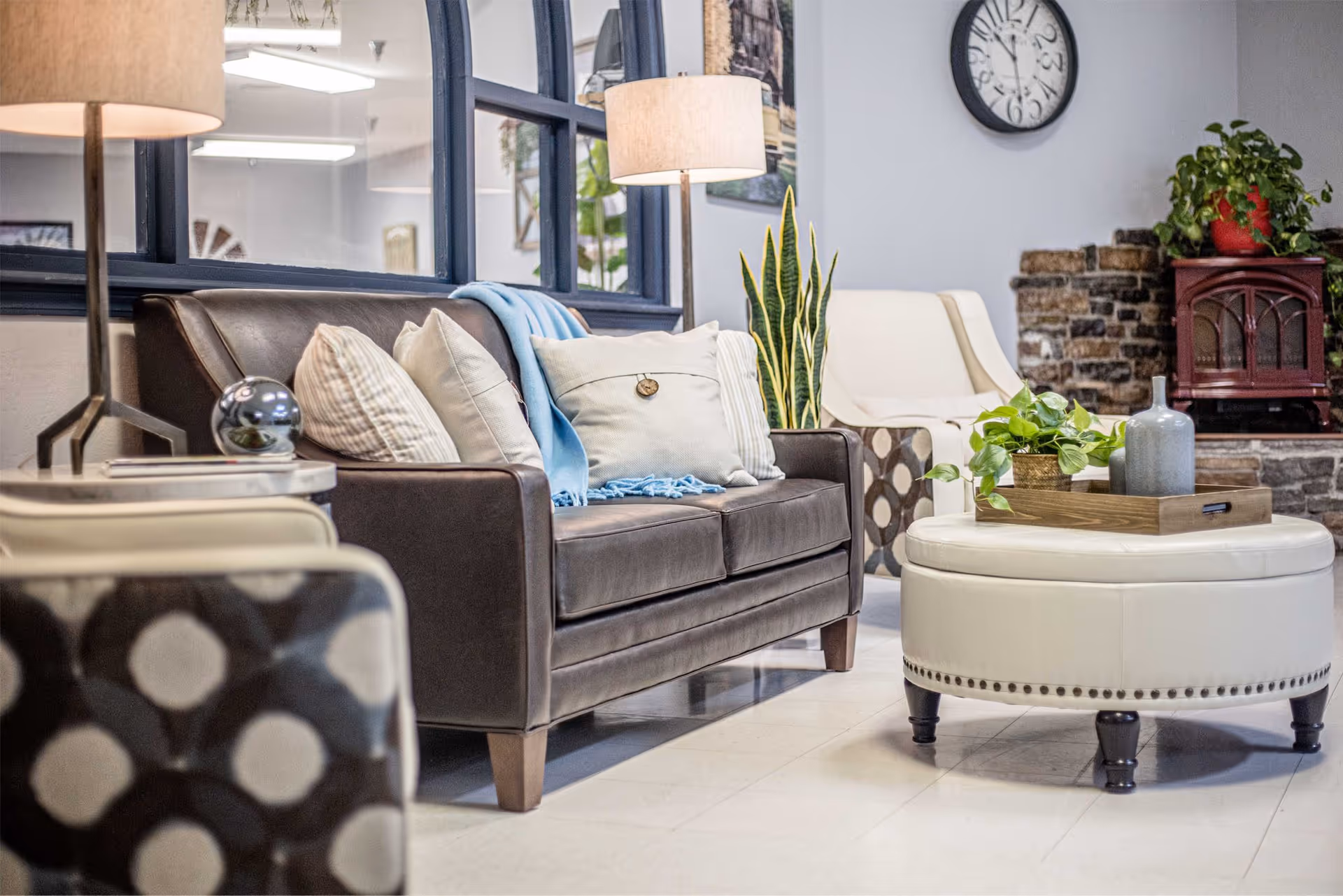 Comfortable lounge area with a dark leather sofa, patterned chairs, round ottoman with a decorative tray, lamps and plants.