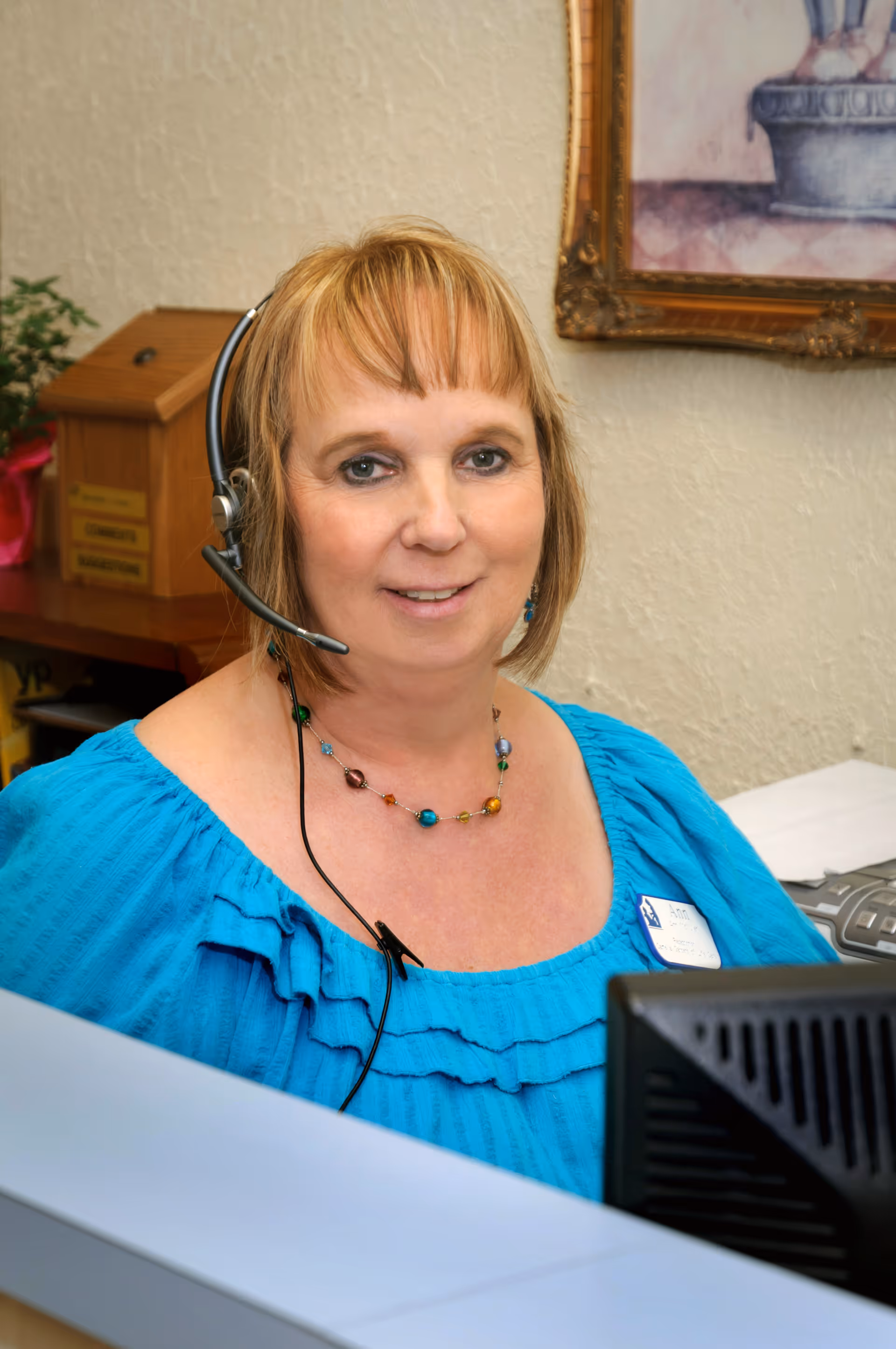 A woman wearing a blue blouse and a colorful beaded necklace is sitting at a reception desk with a headset on, smiling at the camera. Behind her is a wooden box and a framed picture on the wall.