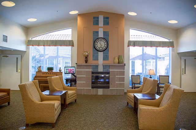 A cozy living room area in a senior living facility with four beige armchairs arranged around two wooden side tables. The room features a central fireplace with a large clock above it and decorative vases on the mantel. Large windows with valances allow natural light to fill the space, and there is a piano and a computer desk visible near the windows.