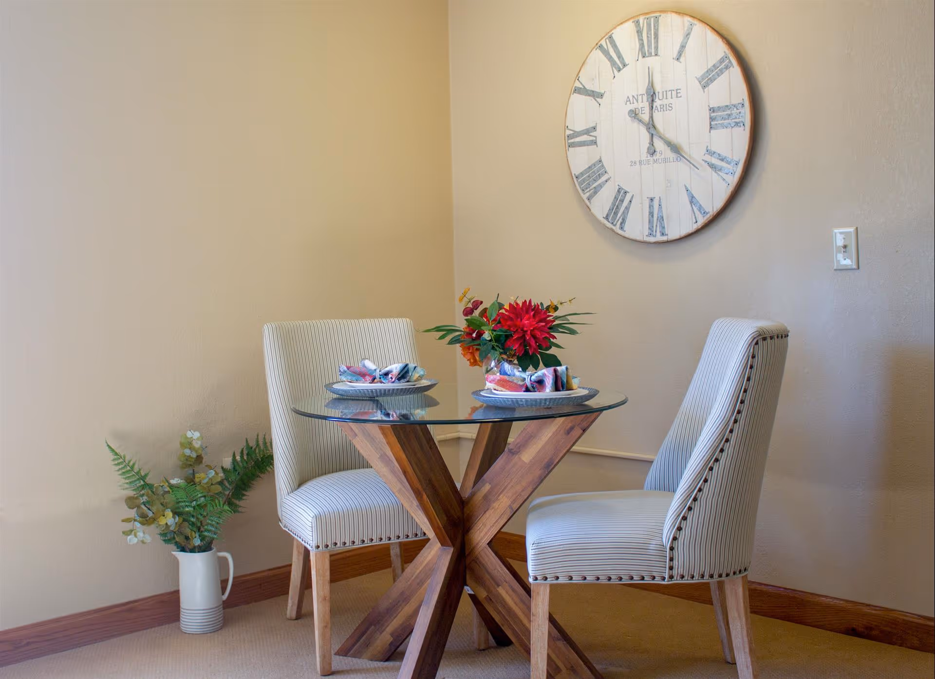 A small dining area with a round glass-top table supported by a wooden X-shaped base, two upholstered chairs with striped fabric and nailhead trim, a vase with red and orange flowers on the table, a large round wall clock with Roman numerals, and a white pitcher with green foliage placed on the floor in the corner.