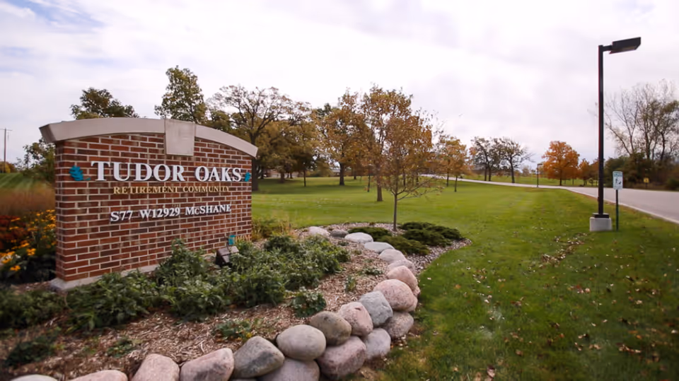 Brick sign for Tudor Oaks Retirement Community surrounded by landscaping with rocks and plants, with a road and trees in the background under a cloudy sky.