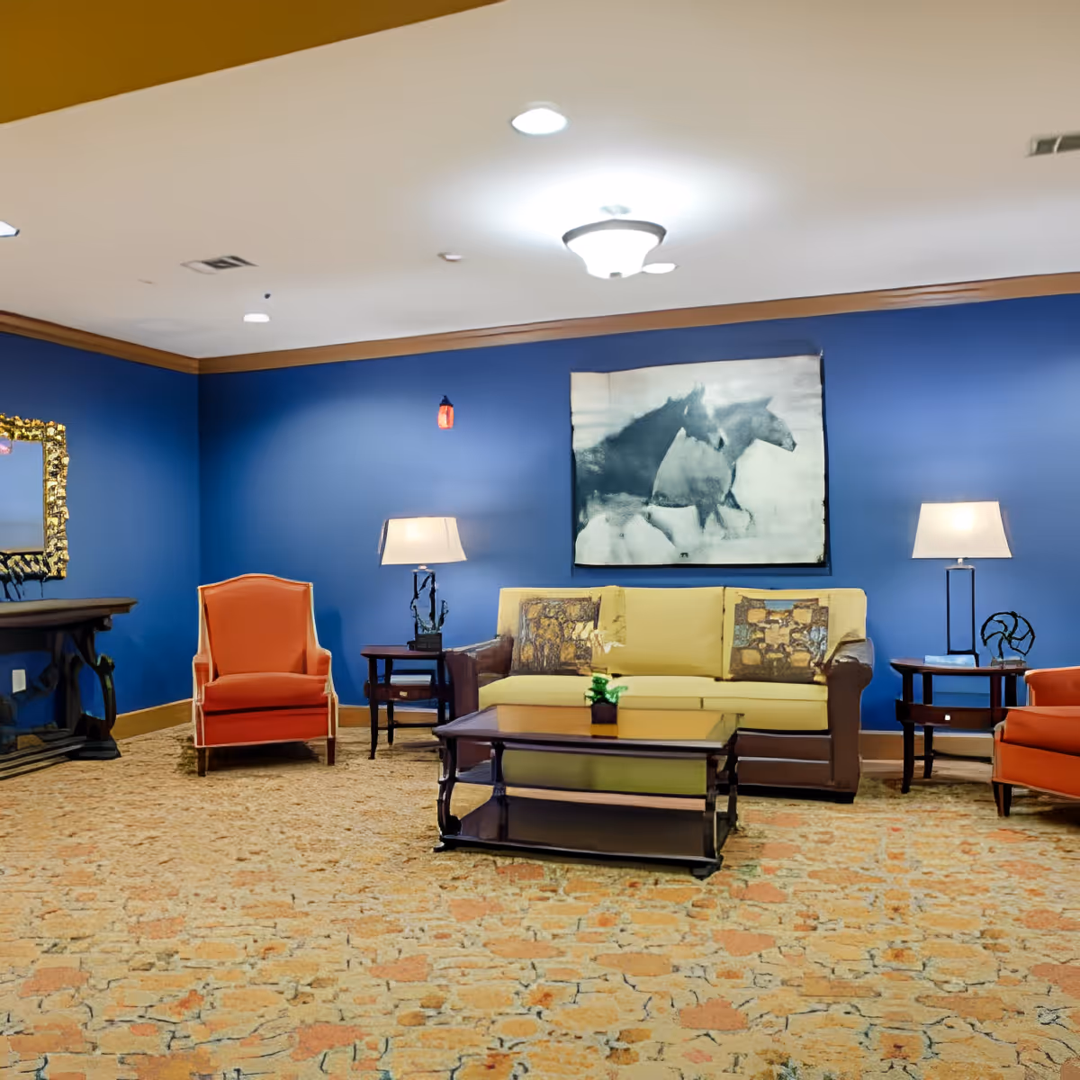 A cozy living room with a yellow sofa and two orange armchairs. The walls are painted blue with wooden trim. There are two side tables with lamps on either side of the sofa, a coffee table in front, and a framed black and white picture of horses hanging on the wall. The floor is covered with a patterned carpet.