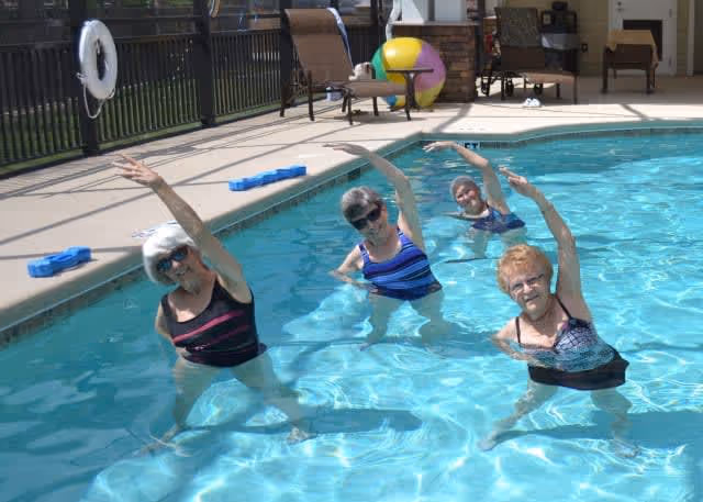 Four elderly women in swimsuits participating in a water exercise class in an outdoor swimming pool, each raising one arm above their heads while standing in the water.
