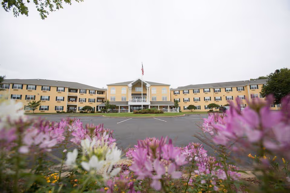 Front view of a three-story yellow senior living building with an American flag and pink flowers in the foreground.