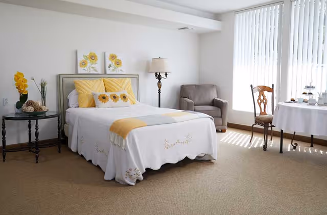 A bright and cozy bedroom with a neatly made bed featuring white and yellow bedding, two sunflower paintings above the headboard, a small round side table with a yellow flower arrangement, a floor lamp, a comfortable armchair, and a table with a chair near a window with vertical blinds letting in natural light.