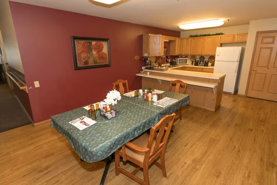 Interior view of a senior living facility dining area with a table covered by a green tablecloth, set with placemats, condiments, and a flower centerpiece. The dining area is adjacent to a kitchen with wooden cabinets, a white refrigerator, microwave, and countertop. The walls are painted red and beige, and there is a framed floral painting on the wall.
