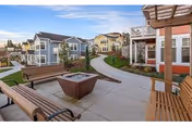 Outdoor seating area with wooden benches arranged around a square fire pit on a paved patio. In the background, there are multiple two-story residential buildings with balconies and well-maintained landscaping including grass and small trees under a clear sky.