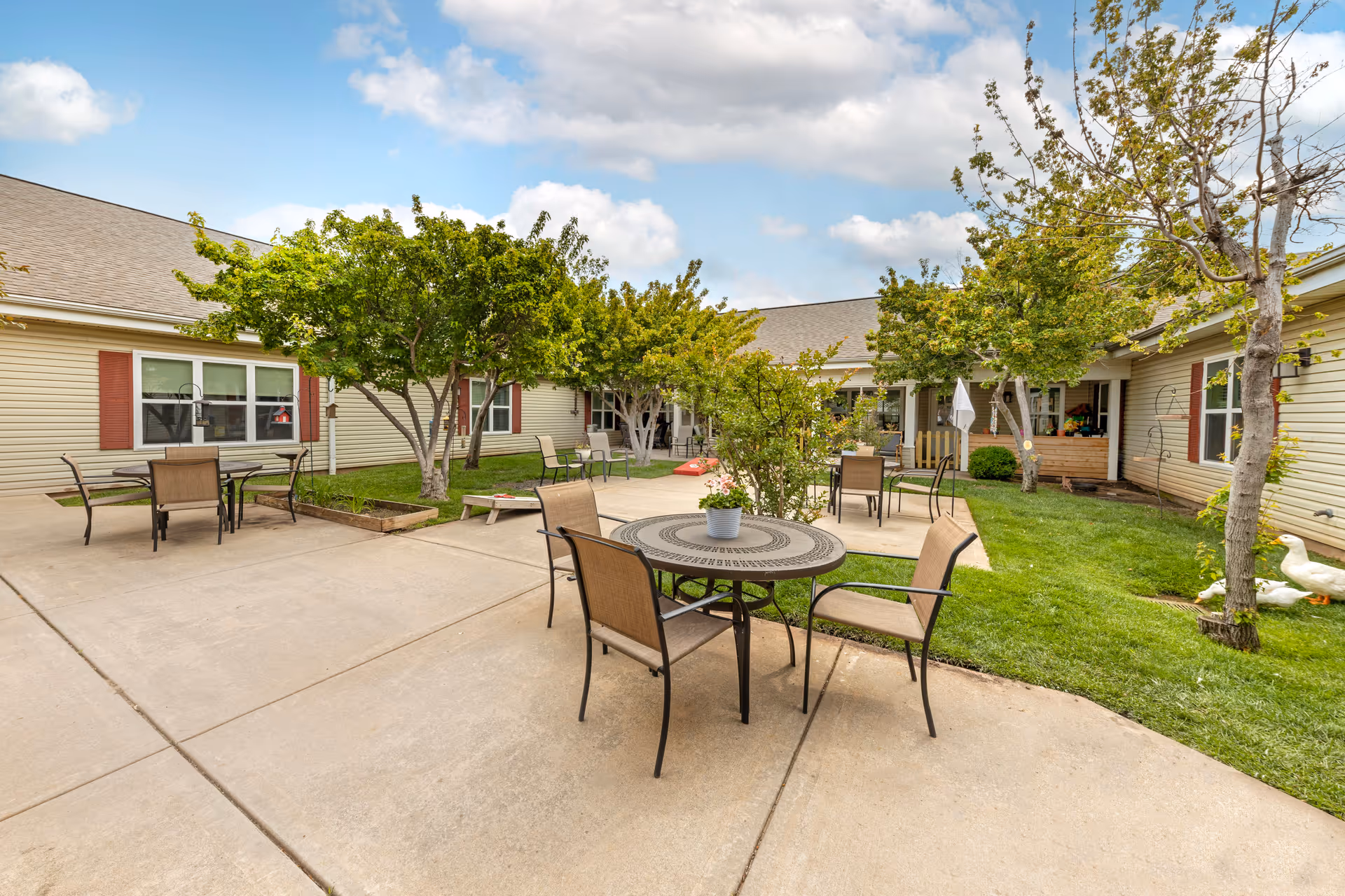 Outdoor courtyard area at Brookdale Oklahoma City Southwest featuring several round tables with chairs on a concrete patio, surrounded by green grass, trees, and beige buildings with red shutters under a partly cloudy sky.