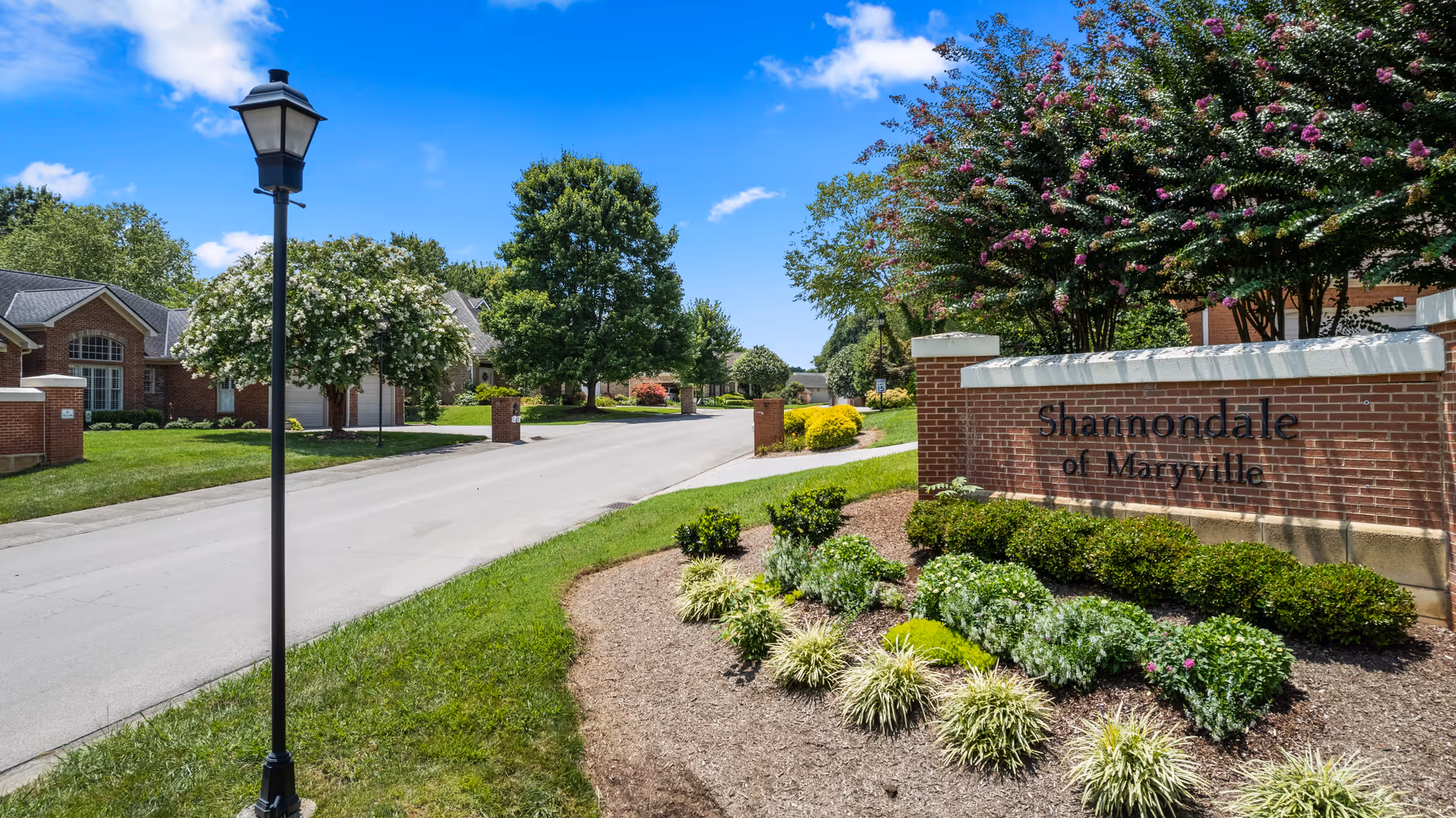 Entrance to Shannondale of Maryville senior living community with a brick sign surrounded by landscaped bushes and flowers, a street lined with trees and houses under a blue sky with some clouds.