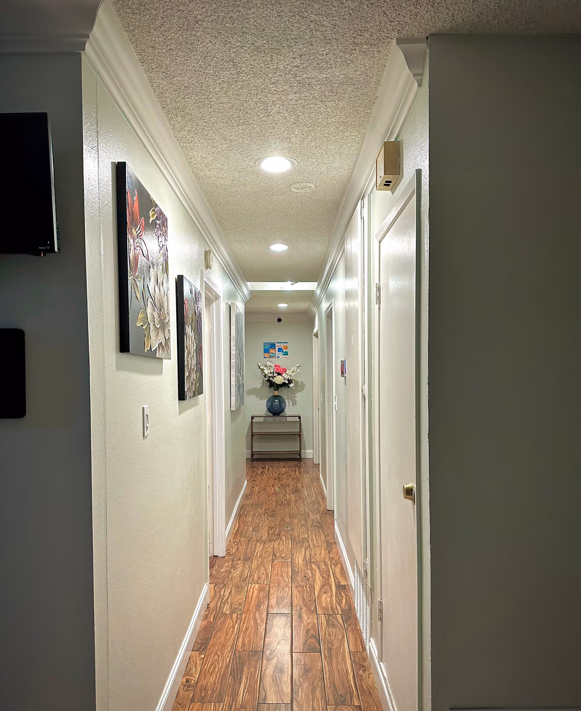 A well-lit indoor hallway with wooden flooring and white walls. The hallway has several closed doors on both sides and decorative floral paintings on the left wall. At the end of the hallway, there is a small table with a blue vase holding a bouquet of flowers. The ceiling has recessed lighting fixtures.