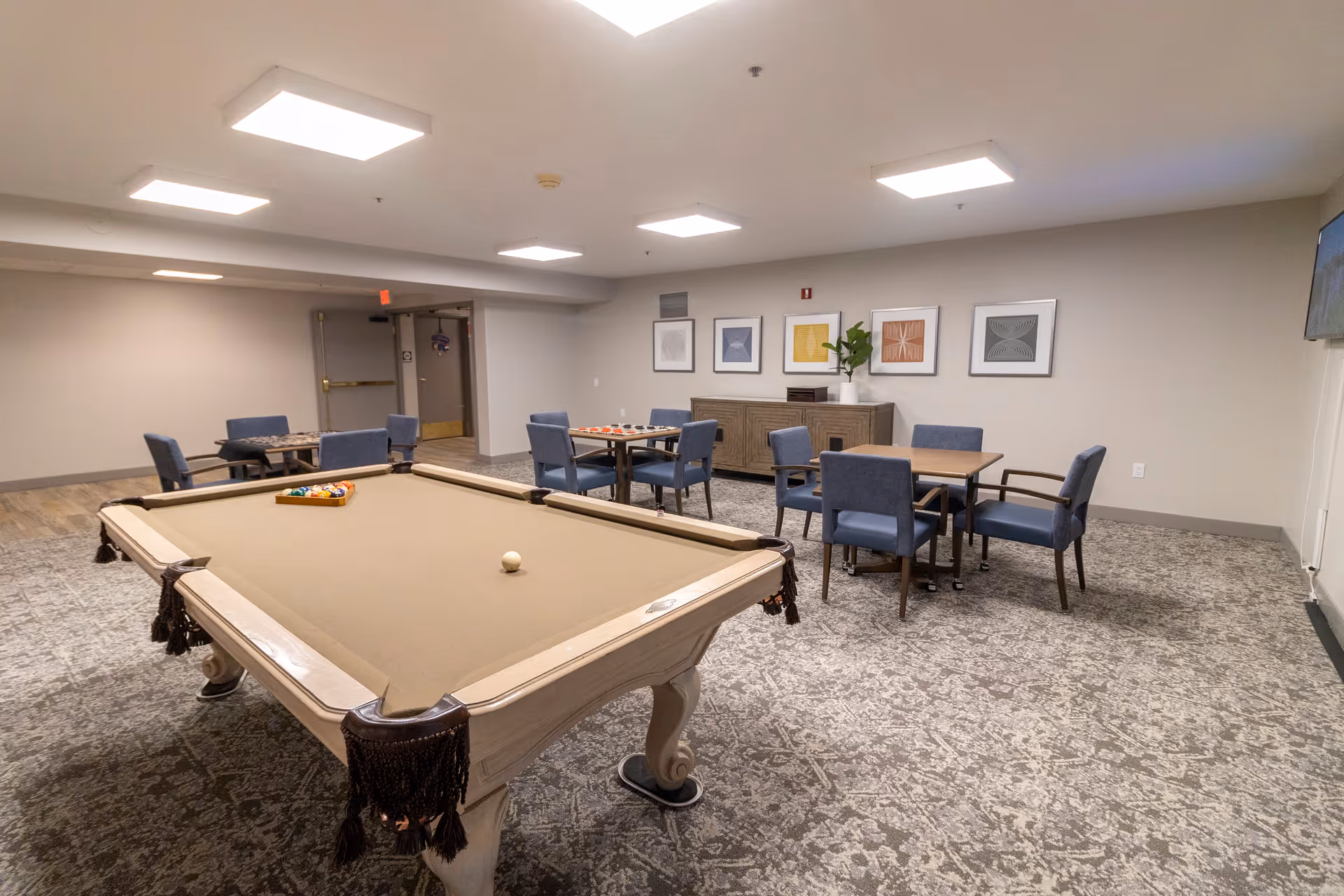 A spacious recreational room with a beige pool table in the foreground, set with billiard balls. In the background, there are several tables with blue upholstered chairs arranged around them. The walls are decorated with framed abstract artwork, and a sideboard with a plant on top is visible. The room has bright ceiling lights and a patterned carpeted floor.