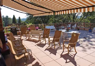 Shaded outdoor terrace with chairs and tables under a striped awning overlooking trees and distant hills.