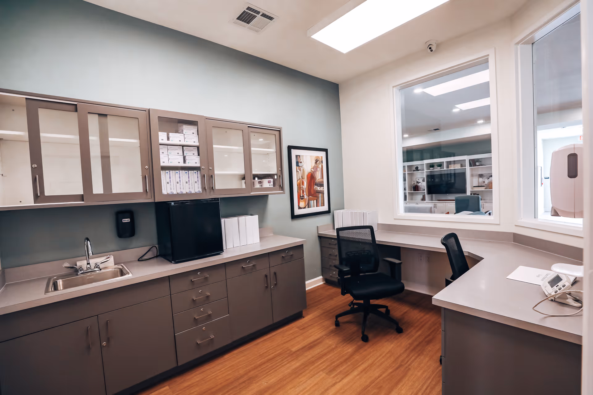 An office or nurse station area with gray cabinets, a small sink, a black mini fridge, office chairs, and a large window looking into another room. The walls are painted light green and white, and there is a framed artwork on the wall. The floor is wood.