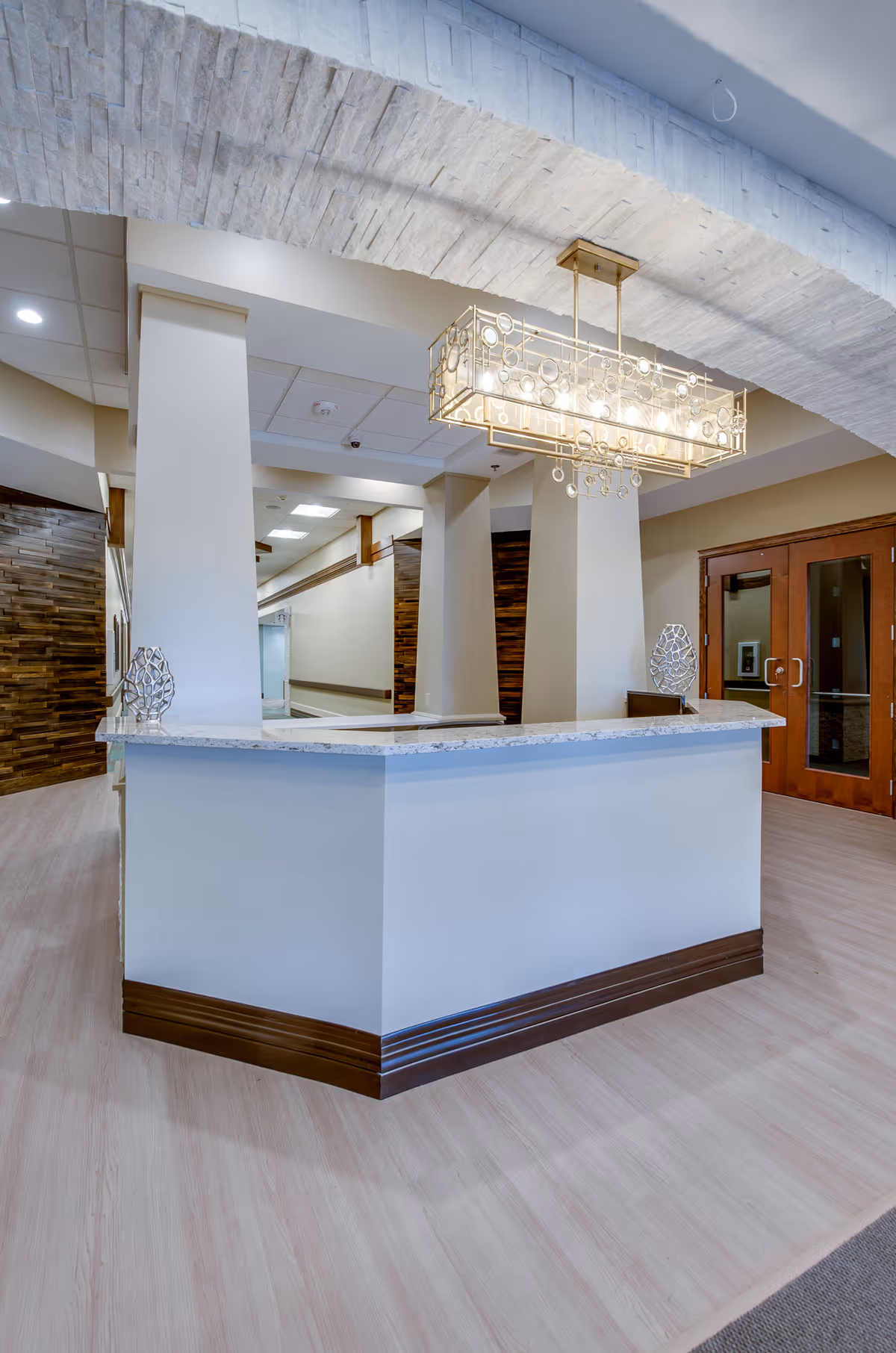 A modern reception desk area inside a facility with a white marble countertop, two decorative sculptures on the counter, a large rectangular chandelier with circular accents hanging from the ceiling, and wooden double doors in the background.