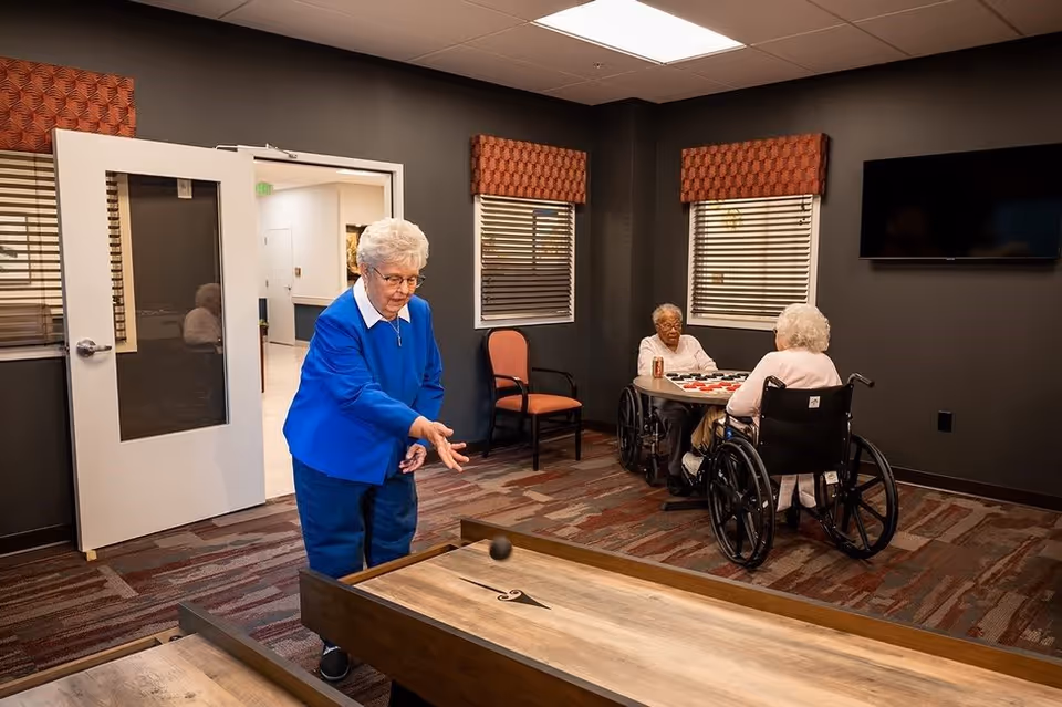 An elderly woman in a blue sweater is playing a tabletop shuffleboard game in a room with dark walls and patterned carpet. In the background, two elderly women in wheelchairs are sitting at a table playing checkers. The room has windows with blinds and a wall-mounted TV.