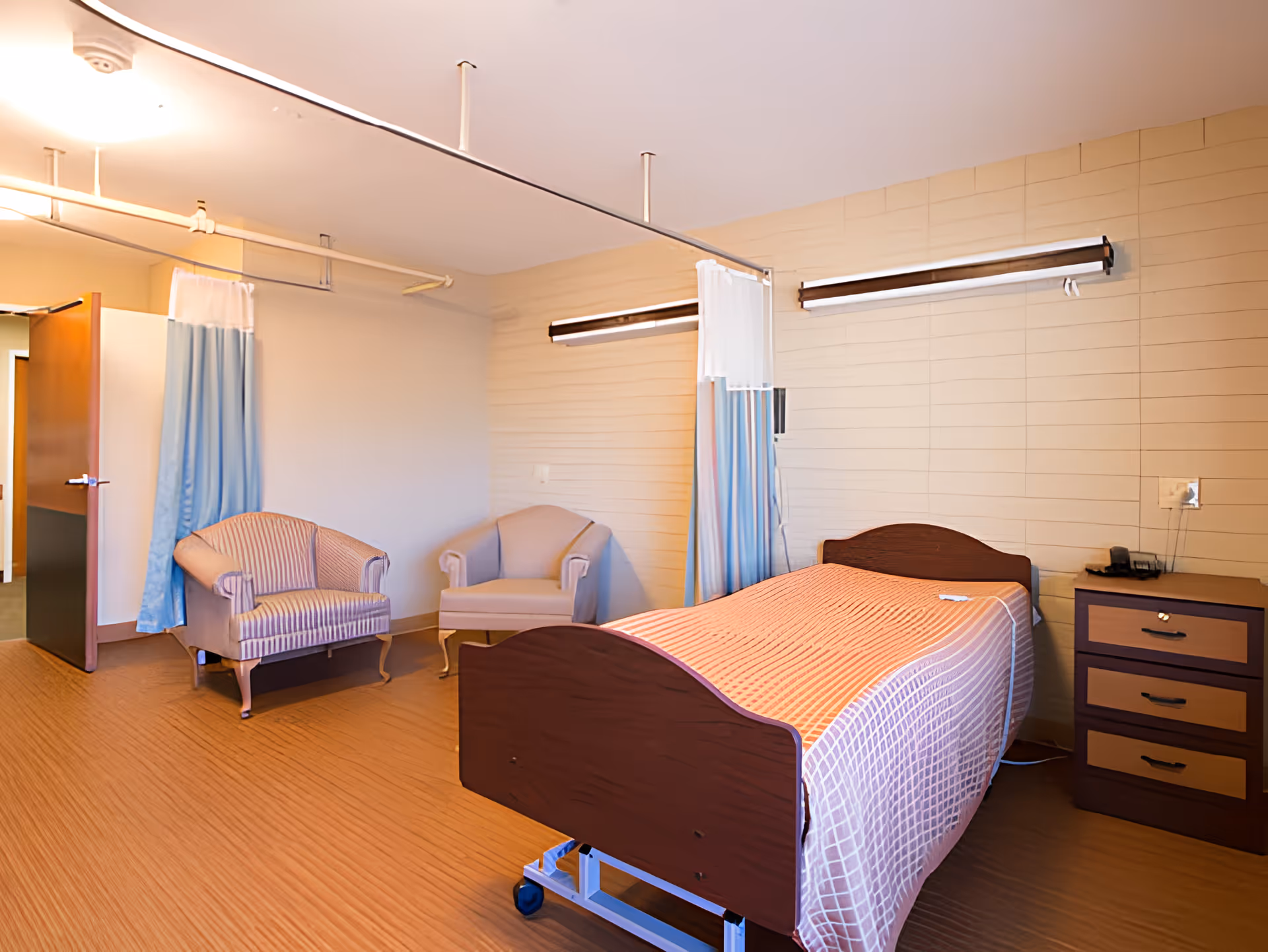 A patient room in a healthcare facility featuring a single hospital bed with a wooden headboard and footboard, a bedside table with drawers, two upholstered armchairs, and privacy curtains hanging from ceiling-mounted tracks. The room has light-colored walls and wood-patterned flooring.