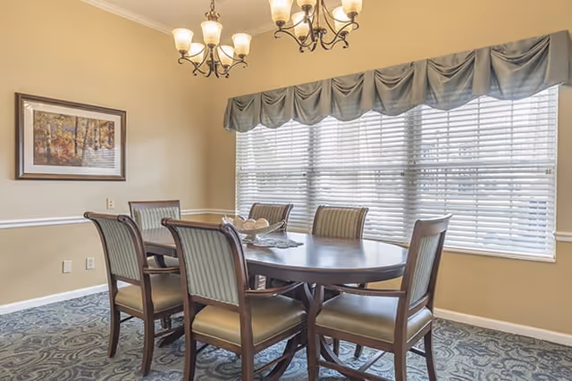 A dining room with a wooden oval table surrounded by six cushioned chairs. The room has a large window with white blinds and a blue valance. Two chandeliers with multiple lights hang from the ceiling. A framed picture of a forest scene is on the beige wall.