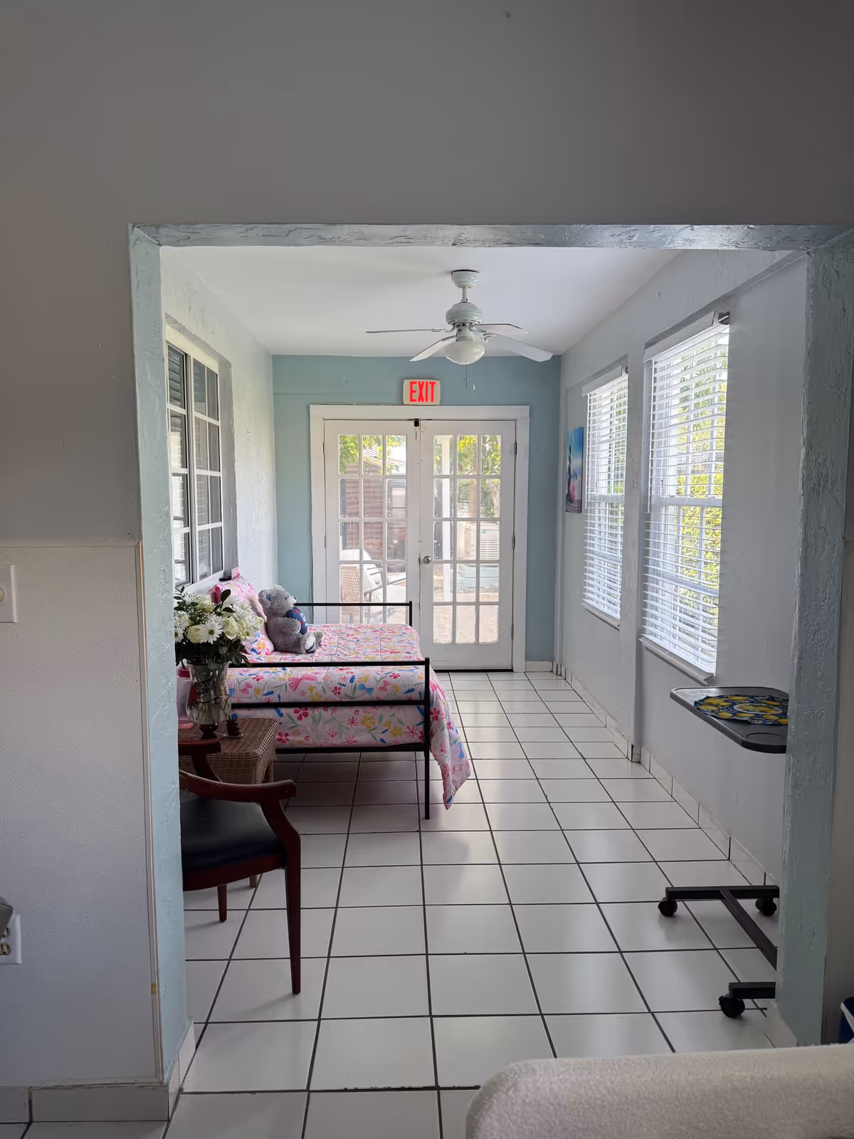 A bright room with white tiled floor and light blue walls featuring a daybed with floral bedding and a teddy bear on it. There is a wooden chair with a black seat cushion and a small table with a vase of flowers next to the daybed. The room has large windows with white blinds on the right side and glass double doors with an exit sign above them at the far end. A ceiling fan with a light is mounted on the white ceiling.