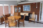 A cozy indoor seating area with a round wooden table surrounded by four wooden chairs. There is a wooden rocking chair to the right and a glass-front cabinet against a brick wall in the background. A framed picture hangs above a black piano or cabinet, and large windows on the left let in natural light.