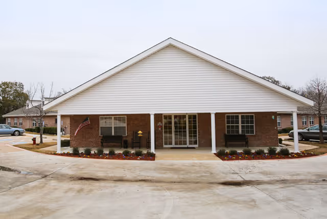 Front entrance of a single-story brick building with a covered porch, benches, an American flag, and landscaped flowerbeds.