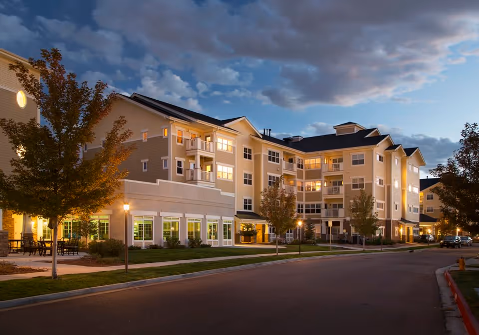 Exterior view of a multi-story senior living facility building at dusk with lights glowing from the windows. The building has balconies, trees along the sidewalk, and a street in the foreground under a partly cloudy sky.