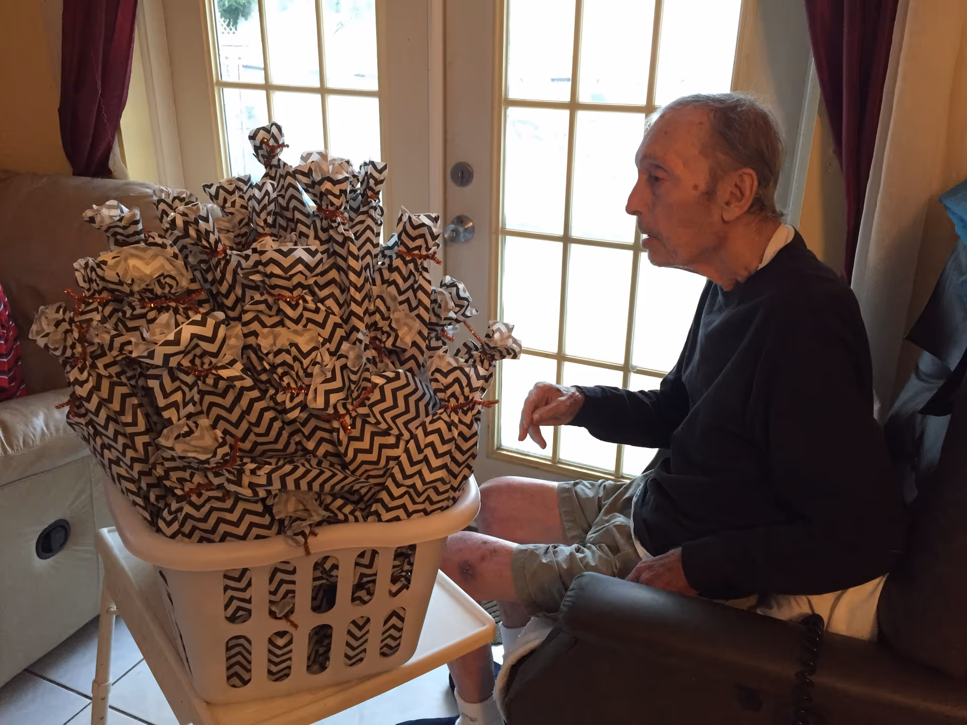 An elderly man sitting in a recliner chair next to a laundry basket filled with many small packages wrapped in black and white zigzag patterned paper tied with orange twist ties, in a room with glass-paneled doors and curtains.