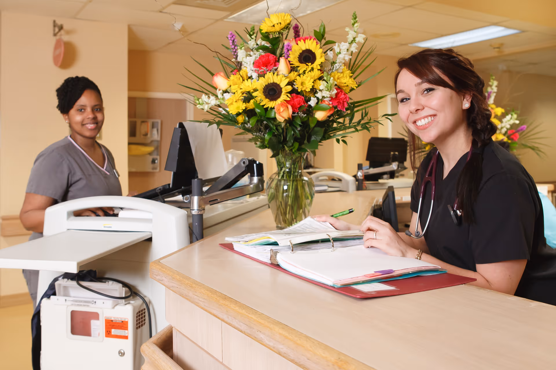 Two healthcare workers smiling at a reception desk in a medical or senior care facility. One woman is standing behind the desk wearing gray scrubs, and the other is sitting at the desk wearing black scrubs with a stethoscope around her neck. A large bouquet of colorful flowers is placed on the desk.