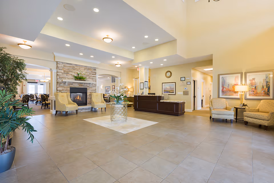 Bright, spacious assisted living lobby with a stone fireplace, seating areas, a reception desk, and decorative plants.