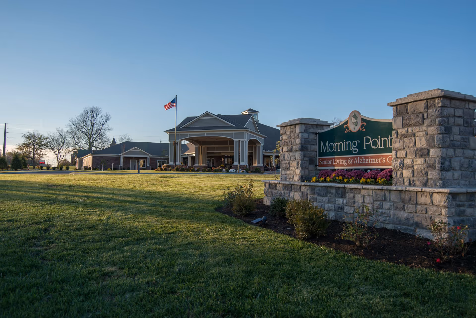Exterior view of Morning Pointe Senior Living & Alzheimer's Center with a large stone sign in the foreground and a building with a covered entrance and an American flag in the background under a clear blue sky.