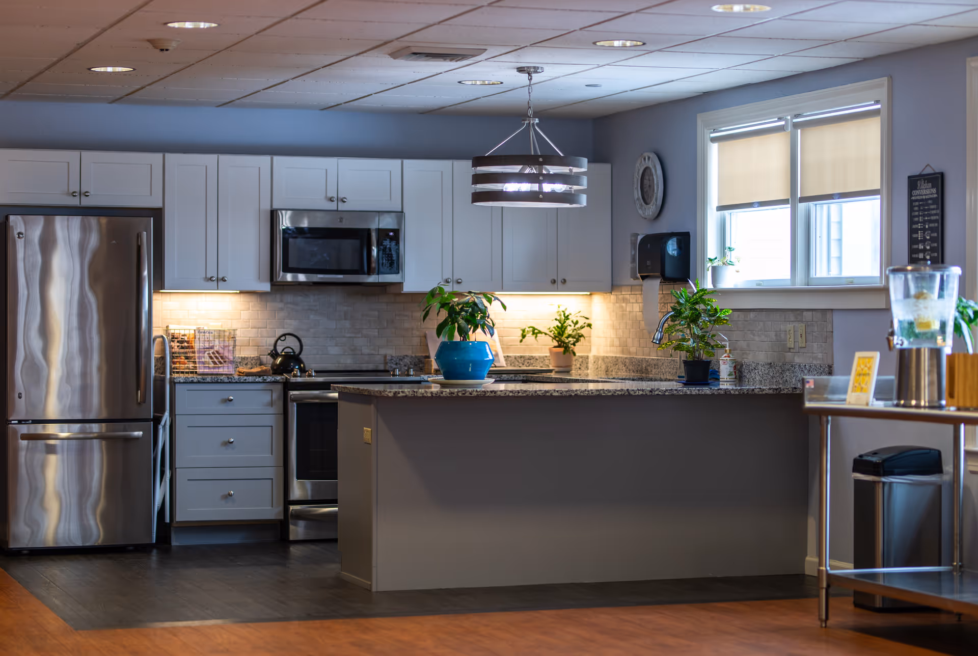 Modern kitchen area with stainless steel refrigerator, microwave, and oven. White cabinets and granite countertops with potted plants on the island and counter. A window with beige blinds lets in natural light. A water dispenser and trash can are visible on the right side.