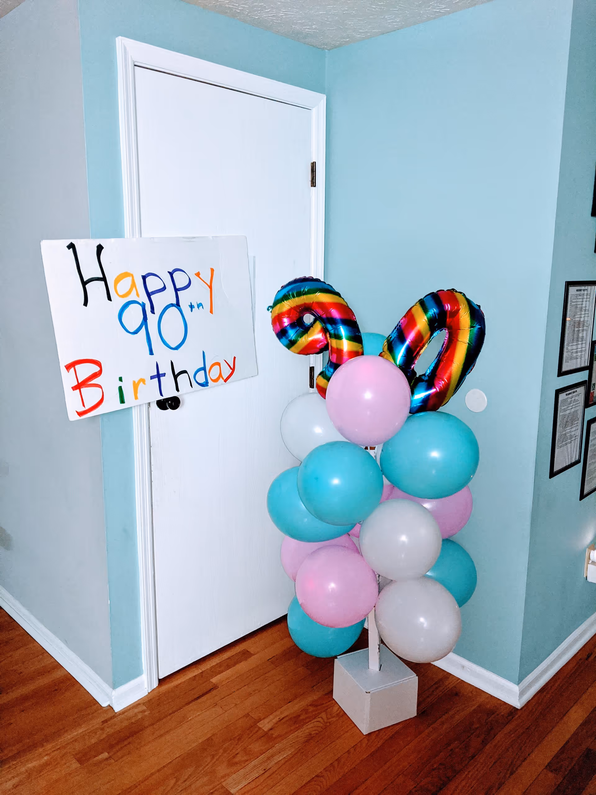 A corner of a room with light blue walls and wooden floor. There is a white door with a colorful handmade sign that reads 'Happy 90th Birthday' attached to the door handle. Next to the door is a balloon arrangement with pastel pink, blue, and white balloons, topped with two large rainbow-colored foil balloons shaped as the number 90.